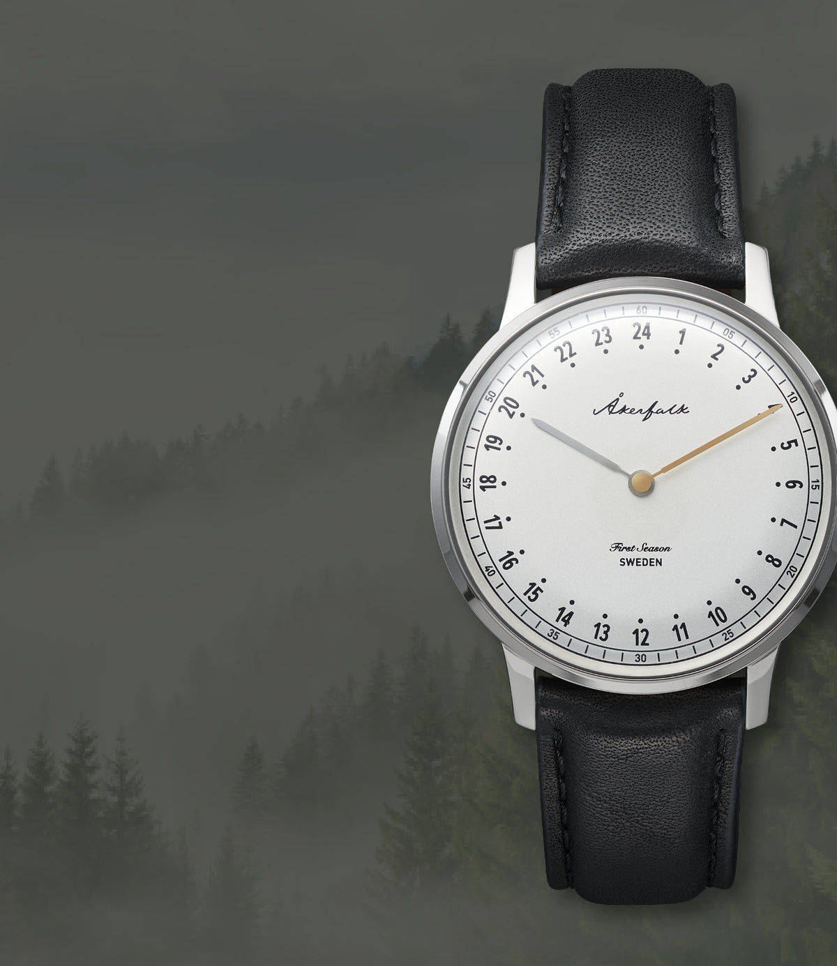A wrist watch with a silver-toned case, white dial, black leather strap, and gold-tone hands, shown against a misty forest background. The brand name 'Aurora' is visible on the dial. 24-hour watch from Akerfalk